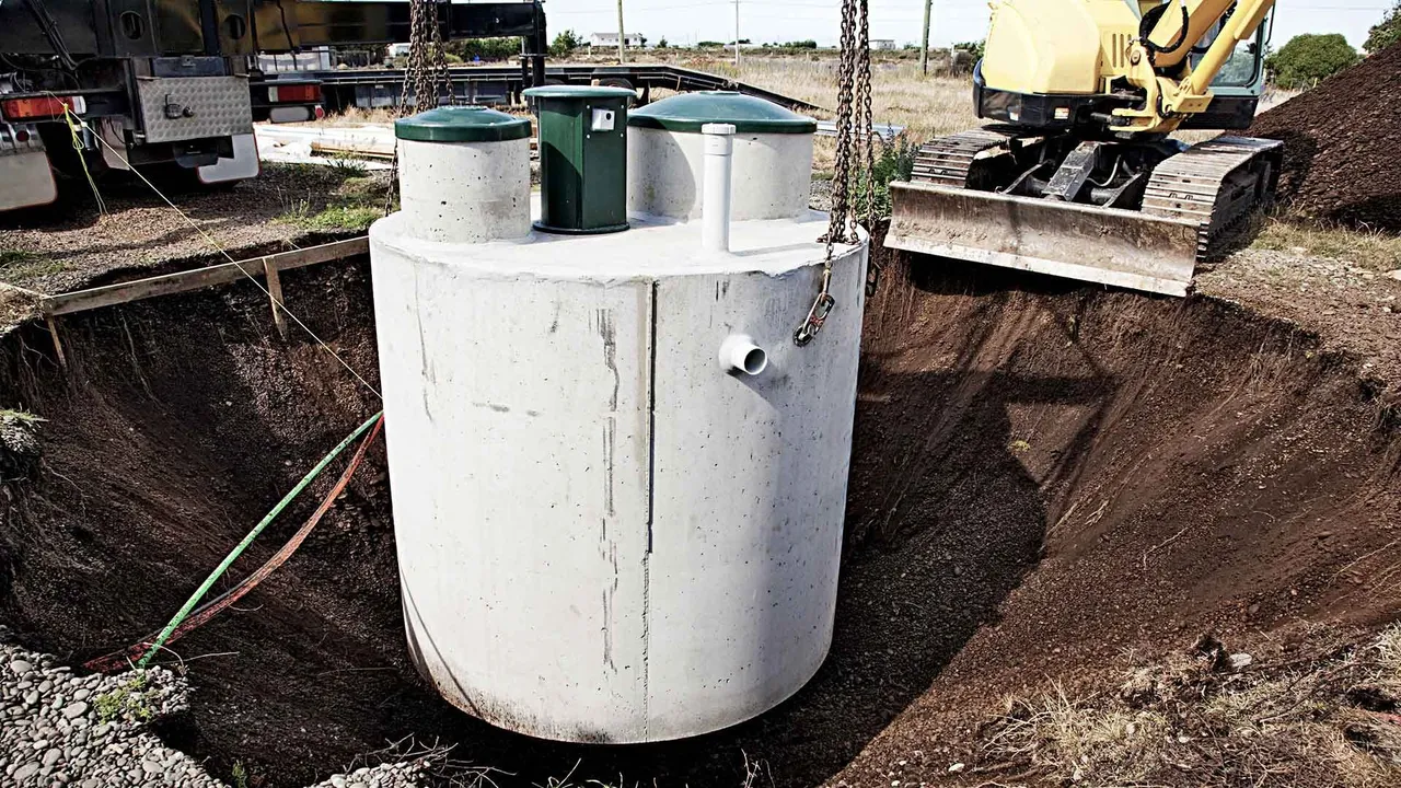 A concrete septic tank being lowered into a large hole by a crane near an excavator.