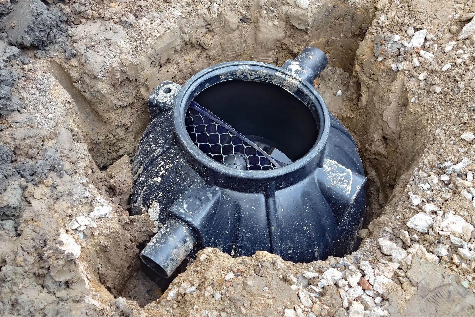 Black plastic water meter, partially buried in a trench, with open lid and connecting pipes. Black plastic water meter, partially buried in a trench, with open lid and connecting pipes.