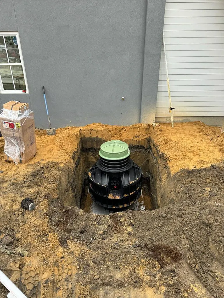 A black septic tank in a square hole in the ground, next to a building and partially covered with dirt.